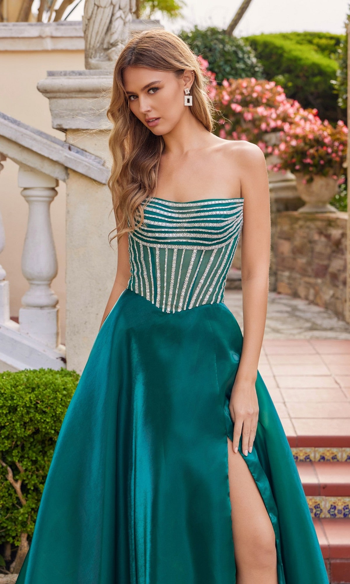A woman stands outdoors by stone steps and blooming flowers, wearing the Long Prom Dress: Juliet JT2663S, featuring a strapless organza design with a beaded sheer bodice and high slit.